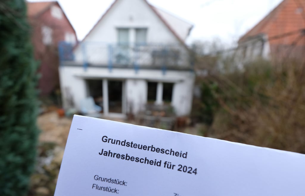 Ein Grundsteuerbescheid für 2024 wird vor einen Computerbildschirm gehalten, auf dem ein Formular der Steuerplattform Elster angezeigt wird.