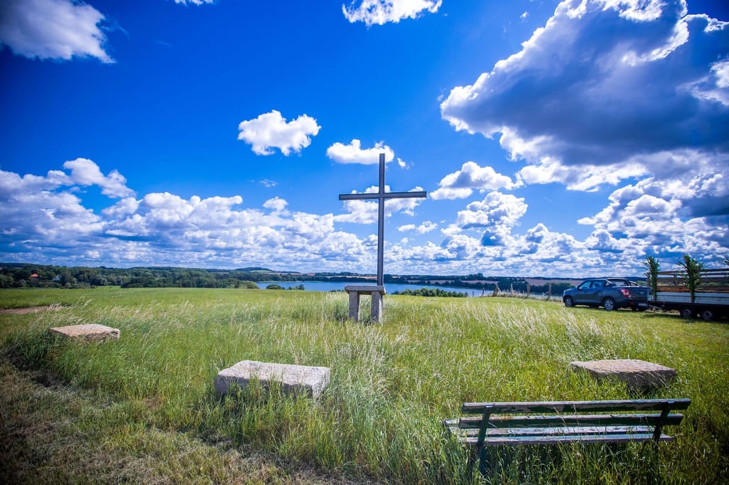Ein großes eisernes Kirchenkreuz steht vor einer imposanten Wolkenkulisse auf dem Bremer Berg über dem Camper See.