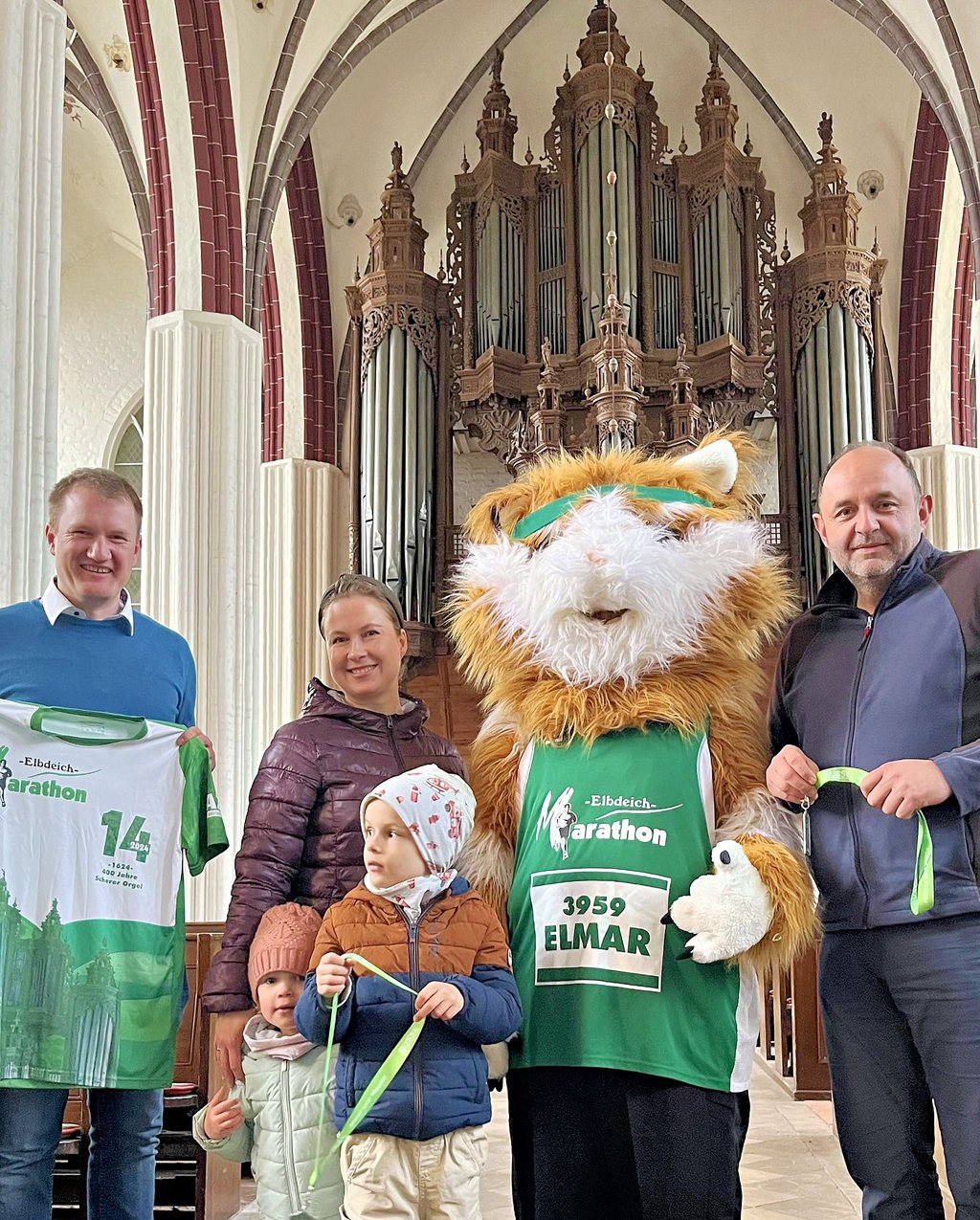 Carsten Birkholz (links) und Thomas Staudt (rechts) vom Tangermünder Elbdeichmarathonverein mit Elmar, dem Vereinsmaskottchen, und Kantorin Olga Minkina in der St. Stephanskirche vor der Schererorgel im Hintergrund.