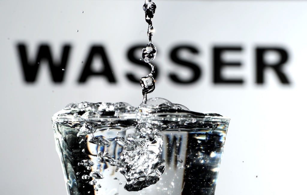Trinkwasser wird vor einem „Wasser“ -Schriftzug in ein Wasserglas gegossen.