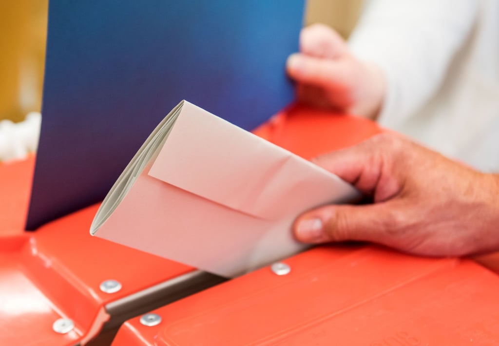 Ein Wahlhelfer steckt in einem Wahllokal den Stimmzettel eines Wählers in eine Wahlurne.