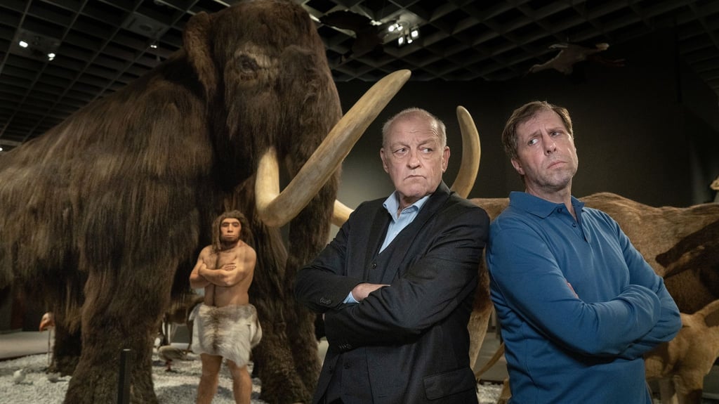 Privatdetektiv Wilsberg (Leonard Lansink) Rücken an Rücken mit Ekki (Oliver Korittke) in einem Raum eines Naturkundemuseums.
