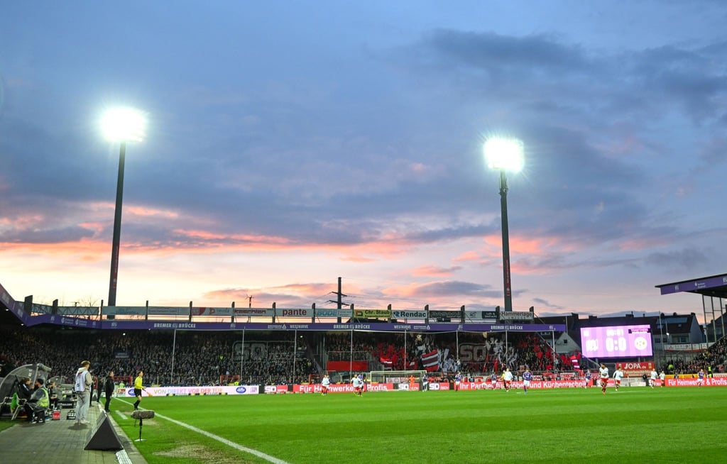 Flutlicht scheint auf das Spielfeld, während im Hintergrund die Sonne untergeht.