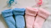 Rosa und blaue Söckchen und dazu die farbigen Nuckel liegen in einem Babybett.