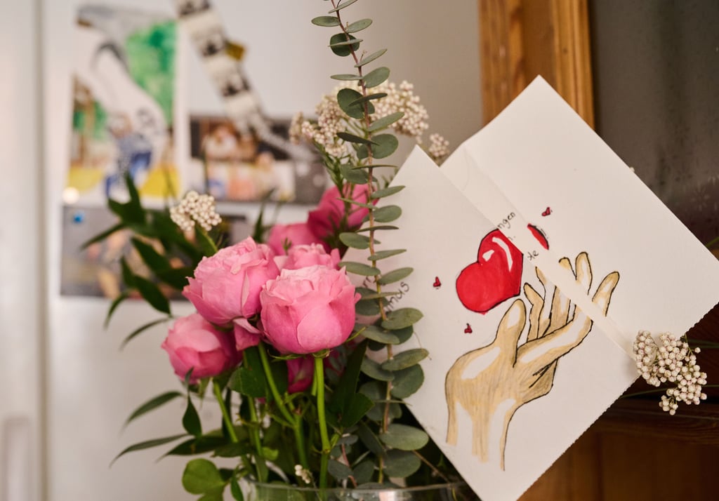 Eine selbstgemalte Karte mit Herz steckt in einem Blumenstrauß, während auf einem Kühlschrank Fotos einer Familie hängen. (Foto: