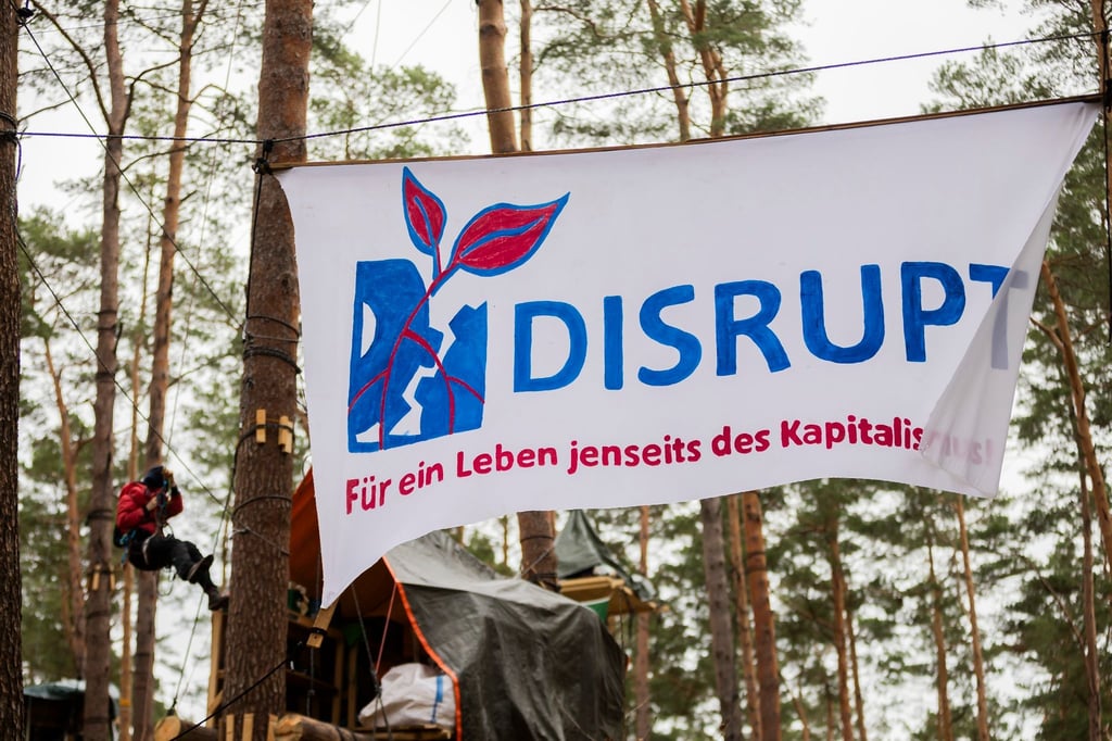 Ein Banner mit der Aufschrift „Disrupt - für ein Leben jenseits des Kapitalismus“ hängt im Camp der Initiative „Tesla stoppen“ in einem Kiefernwald nahe der Tesla-Gigafactory Berlin-Brandenburg zwischen Bäumen.