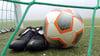 Fußballschuhe und ein Ball liegen hinter einem Tornetz.