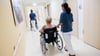 Zwei Pflegerinnen schieben in einem Klinikum Patienten mit Rollstühlen durch einen Flur.