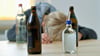 Ein Mann liegt mit seinem Kopf auf einem Tisch, vor ihm stehen leere Bierflaschen und Spirituosen-Flaschen. 