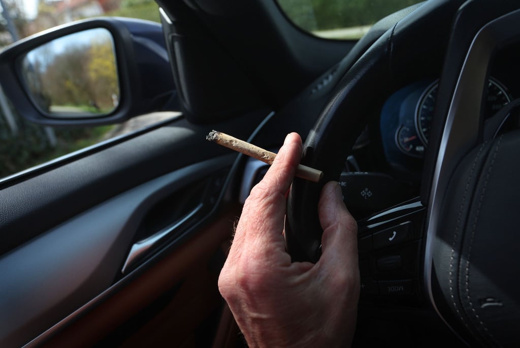 Ein Mann sitzt mit einem Joint zwischen den Fingern am Steuer eines Autos.