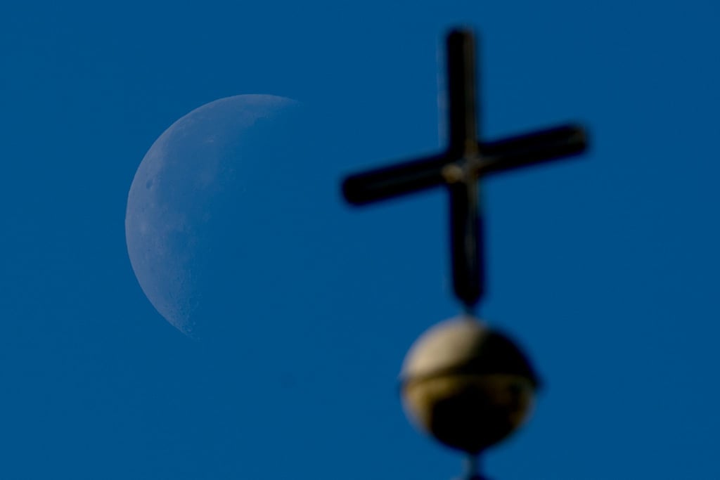 Der abnehmende Viertelmond ist hinter einem Kirchenkreuz zu sehen.