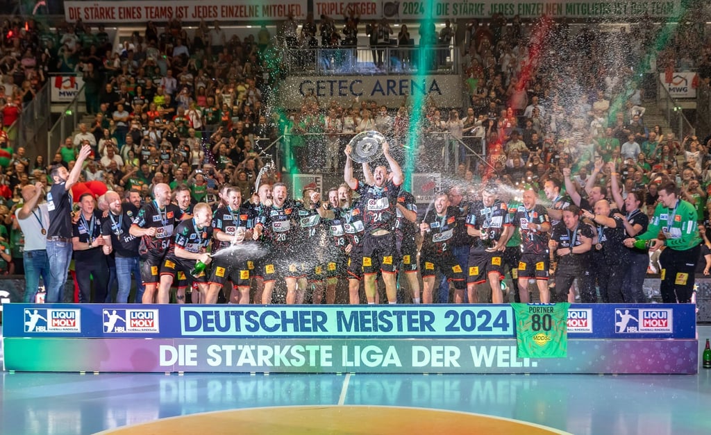 Christian O Sullivan (M) hebt die Meisterschale in die Höhe und jubelt mit Teamkollegen des SC Magdeburg hinter Schriftzug: Deutscher Meister 2024.