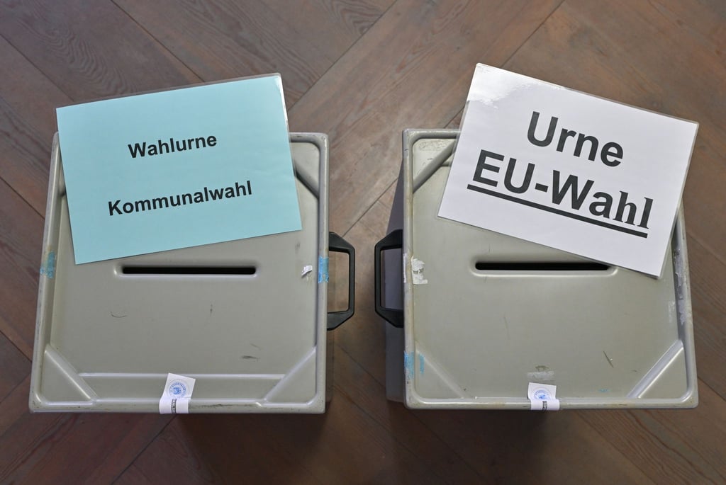 Zettel mit der Aufschrift „Wahlurne Kommunalwahl“ (l) und „Urne EU-Wahl“ liegen auf zwei Wahlurnen in einem Wahllokal.