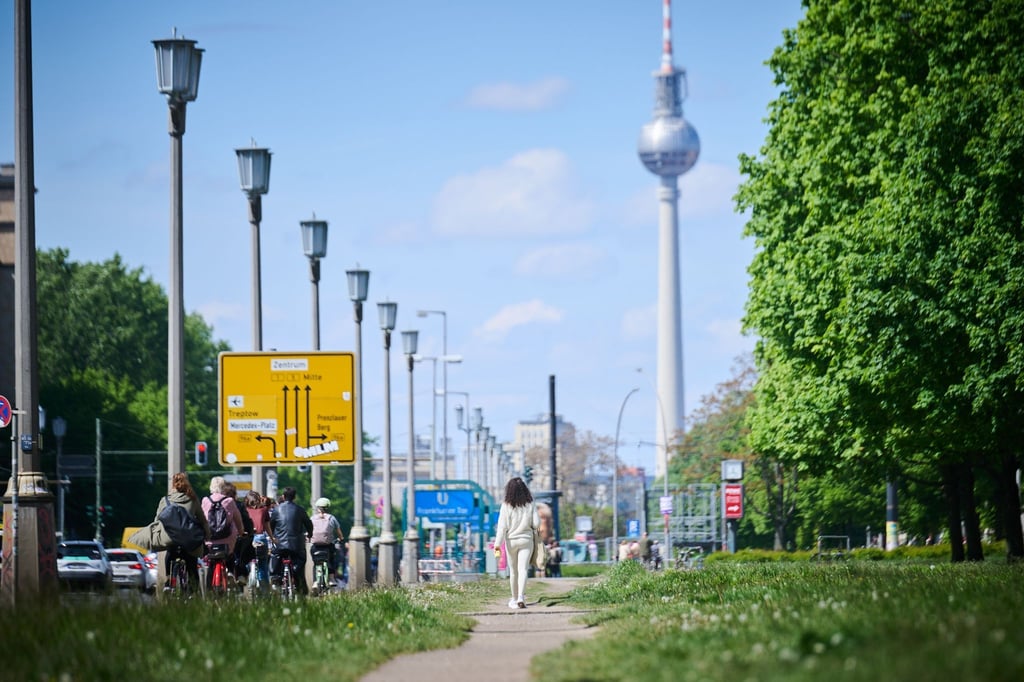 Menschen gehen und fahren an der Frankfurter Allee, während die Sonne auf den Fernsehturm scheint.