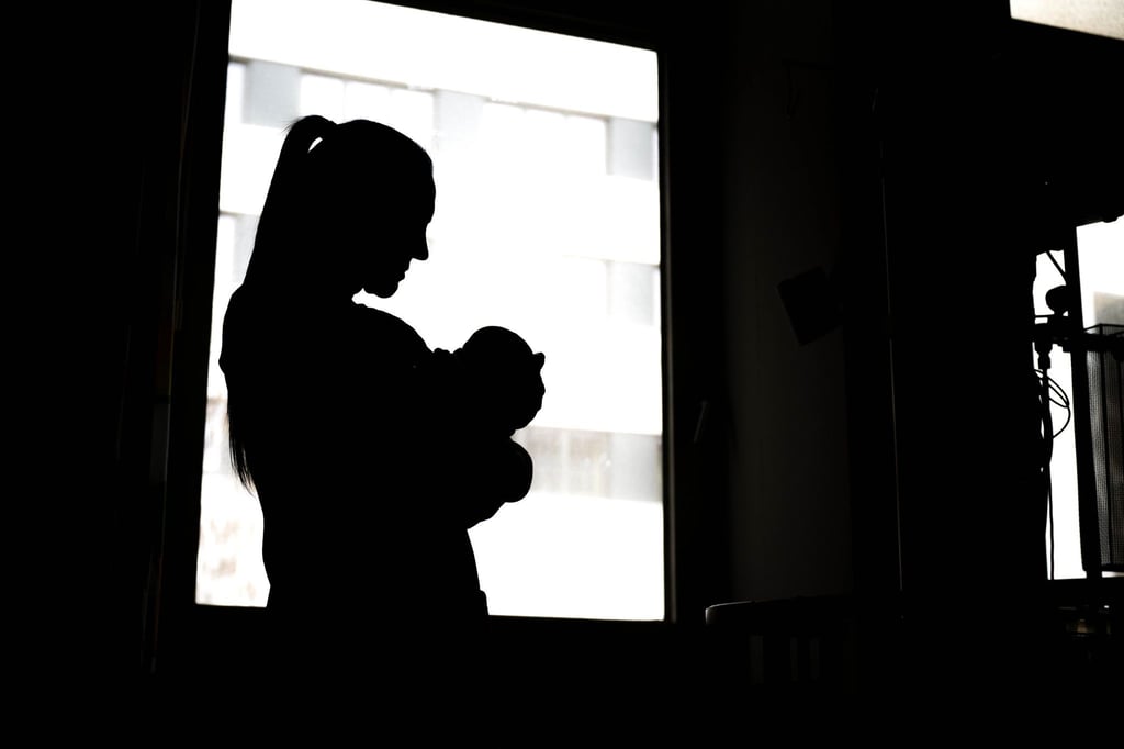Eine Frau steht mit einem Neugeborenen am Fenster einer Klinik.