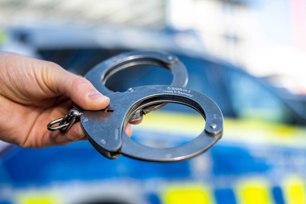 Eine Hand hält Handschellen vor einen Streifenwagen der Polizei.