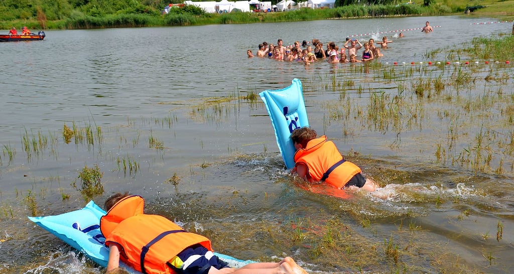 Bela und Lenny starten beim Luftmatratzenrennen für die Jugendfeuerwehren Aschersleben und Winningen. Das Wasser des Güstener Kiesschachts ist warm und klar. Im Hintergrund die Fans und ein Sicherungs-DLRG-Boot.