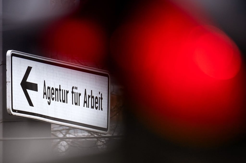 Eine rote Ampel leuchtet vor einem Schild, das auf die Agentur für Arbeit hinweist.