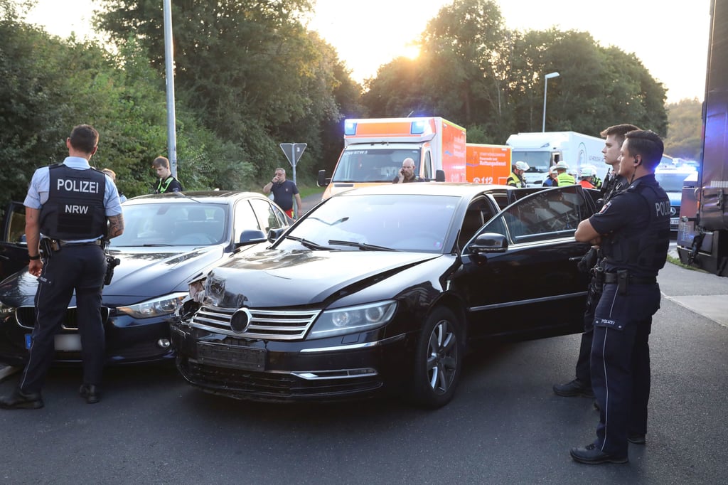 Einsatzkräfte der Polizei und Helfer stehen an einem Tatort an einem Rastplatz an der Autobahn 3 (A3) neben zwei schwarzen Autos.
