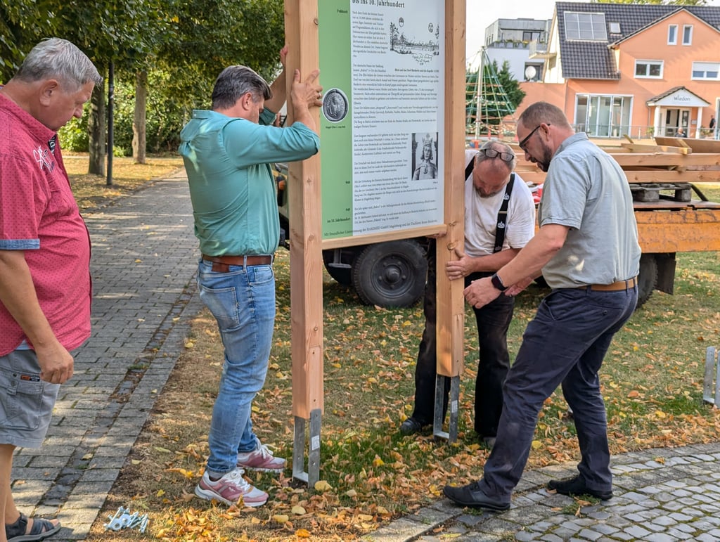 Matthias Weber, Michael Rexin und Kay Gericke passen die gerade vom Transporter abgeladene Infotafel in den Hülsen auf der Kantorwiese ein. Walter Metscher steht bereit zum Mitanfassen dabei.