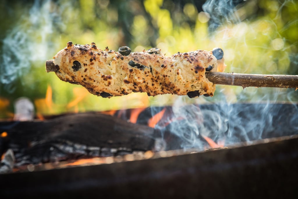 Ein Oliven-Feta-Knoblauch-Hefeteig ist um einen Haselnuss-Stock gewickelt und wird über dem Lagerfeuer gebacken.