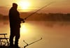 Ein Angler steht am frühen Morgen mit seiner Rute im Licht der aufgehenden Sonne an einem Fischteich. 