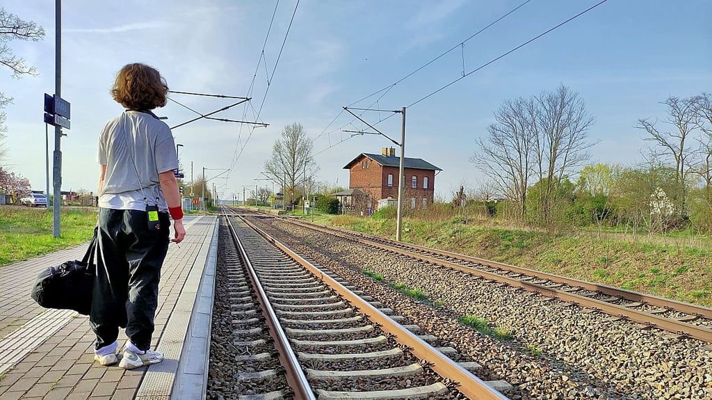Eine Person mit Reisetasche späht am Bahnsteig 1 des Haltepunktes Gnadau nach einem Zug, der aus Richtung Schönebeck erwartet wird.