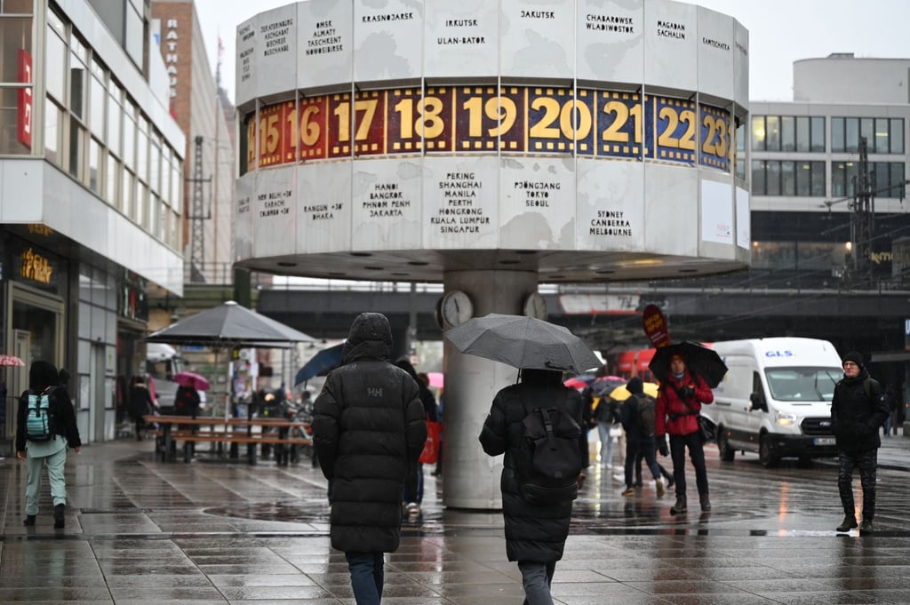 Passanten laufen mit Regenschirmen vor der Weltzeituhr am Alexanderplatz.