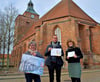 Bettina Huesmann, Gordon Sethge und Gerda Schwander präsentieren stolz den Kalender der Osterburger Perspektiven von Walburga Menke vor der evangelischen Kirche St. Nicolai (Kreis Stendal).