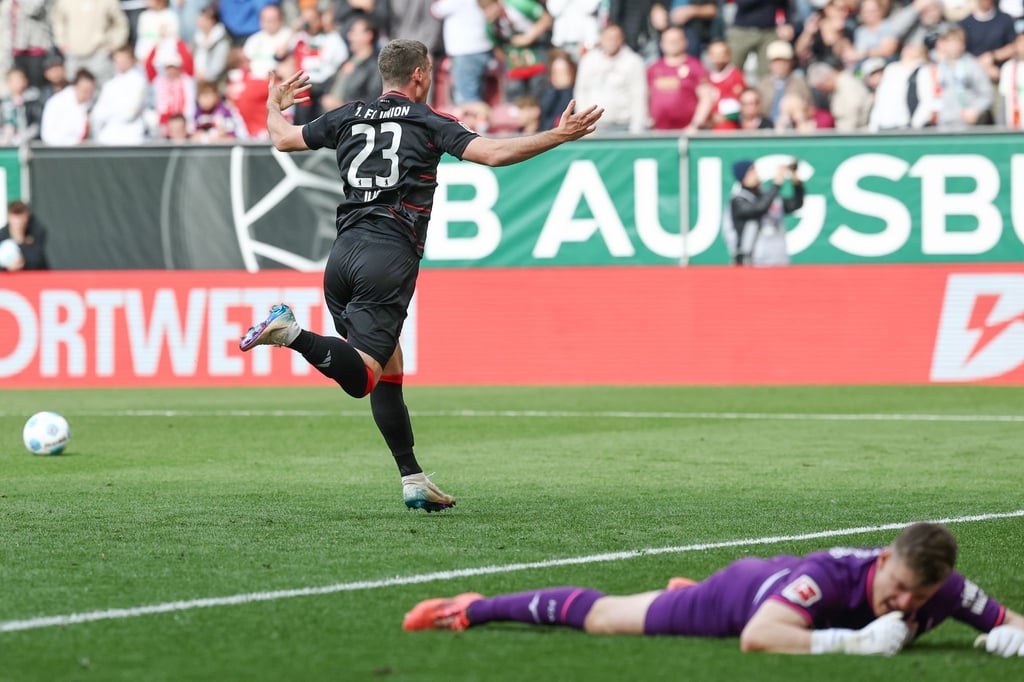 Berlins Matchwinner Andrej Ilic läuft nach seinem Siegtor jubelnd vor den Union-Fanblock. Augsburgs Torwart Finn Fahmen liegt geschlagen am Boden.