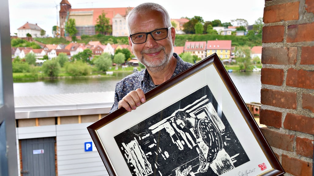Geschäftsführer Andreas Lewerken auf der Schiffswerft in Havelberg mit einem Bild, das ihn in den Anfängen auf dem Kiebitzberg in Kuhlhausen zeigt, wo vor 40 Jahren alles mit dem Herstellen von Holzspielzeug begonnen hat. Der Künstler Henry Korwald aus Kamern hat den Holzschnitt nach einem alten Foto angefertigt.