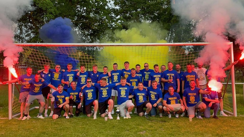 Meister unter sich – Arm in Arm, voller Stolz und mit breitem Grinsen: Spieler der ersten und zweiten Mannschaft feiern den gemeinsamen Aufstieg. Im Hintergrund leuchten blau-gelbe Bengalos – ein emotionaler Moment in Vereinsfarben.