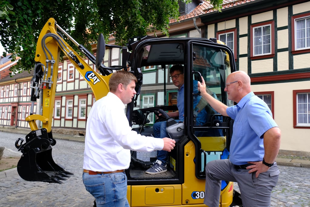 Zeppelin/Cat-Außendienstmitarbeiter Jan Stümpfel erläutert Thomas Schulz, Geschäftsführer der Firma Scheffer Rohrleitungsbau, und Stadtwerke-Chef Steffen Meinecke (von links) die Vorteile des auf der Baustelle in der Wernigeröder Hinterstraße eingesetzten Elektrobaggers.