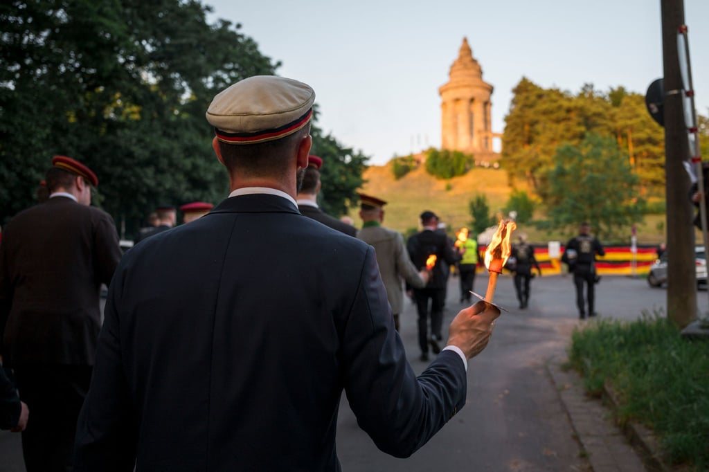 Ein Mitglied einer Burschenschaft hält eine brennende Fackel in der Hand und marschiert auf das Burschenschaftsdenkmal zu.