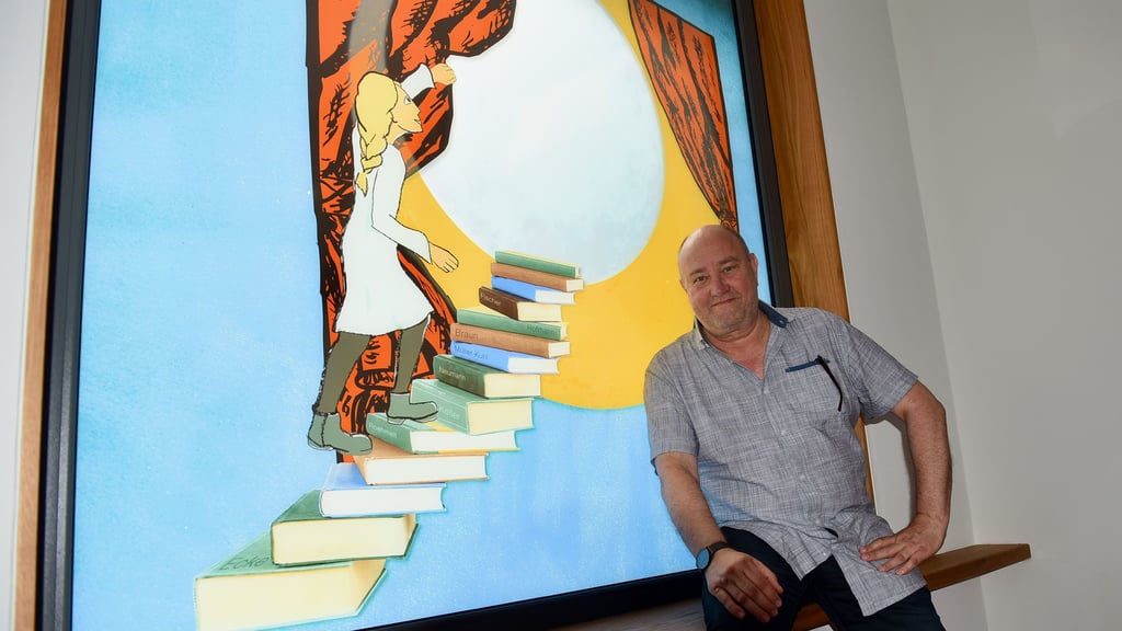 Ulrich Fischer an einem Glasfenster, das eine Grafikerin gestaltet hat. Das Bild zeigt ein Mädchen auf einer Treppe aus Büchern, die mit den Namen der bisherigen Intendanten betitelt sind.
