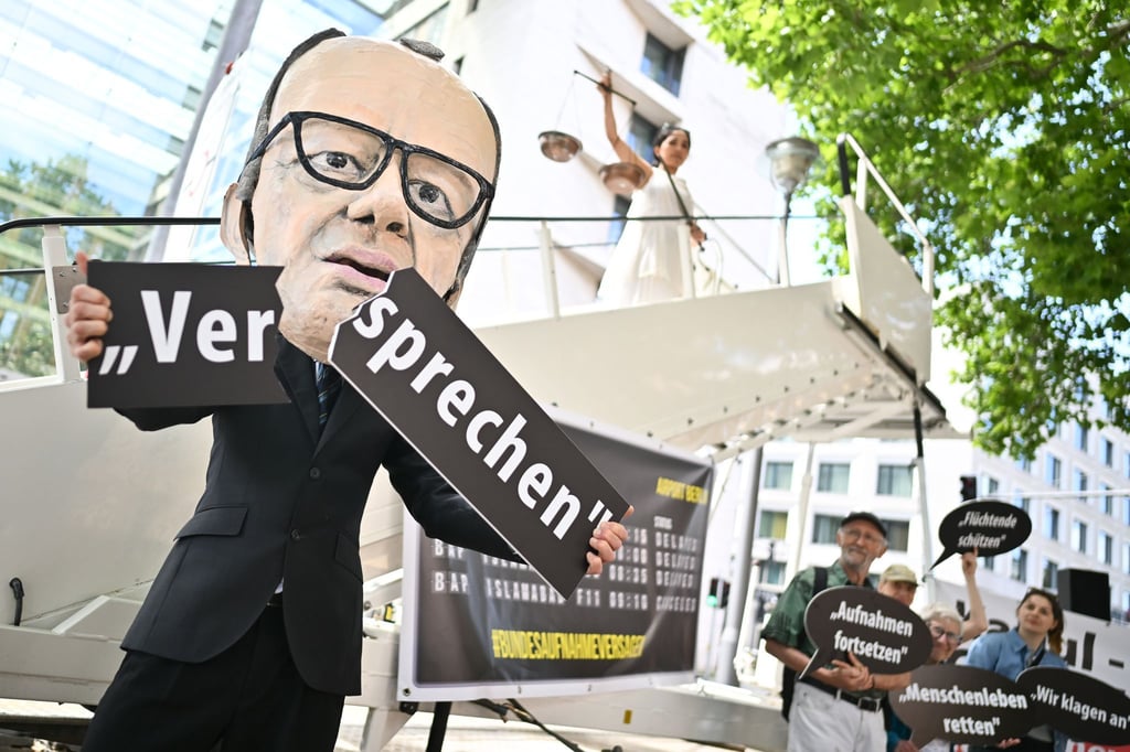 Ein Aktivist mit Merz-Maske zerbricht während einer symbolischen Protestaktion von Kabul Luftbrücke und Campact ein Schild mit der Aufschrift „Versprechen“.