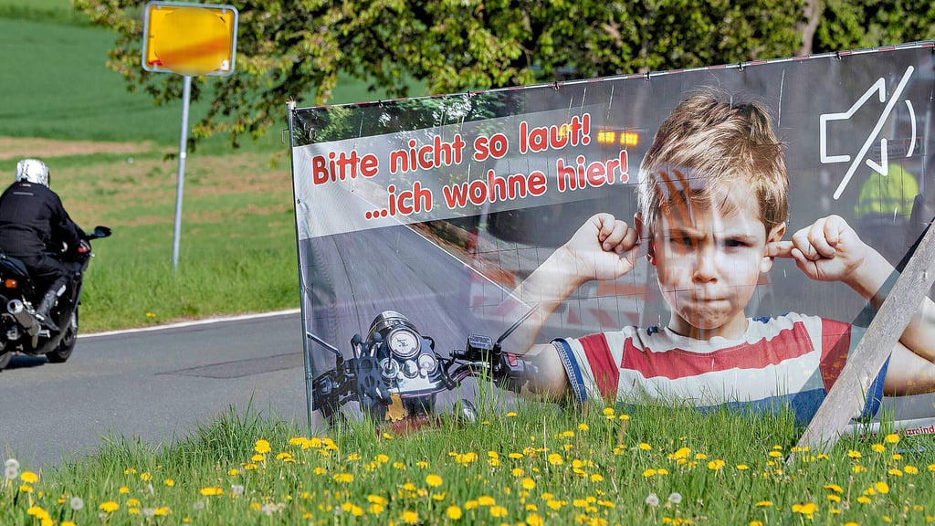 Ein Plakat mit der Aufschrift „Bitte nicht so laut! ... hier wohne ich“ ist an einem Ortsausgang zu sehen, während ein Motorrad daran vorbeifährt.