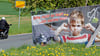 Ein Plakat mit der Aufschrift „Bitte nicht so laut! ... hier wohne ich“ ist an einem Ortsausgang  zu sehen, während ein Motorrad daran vorbeifährt. 