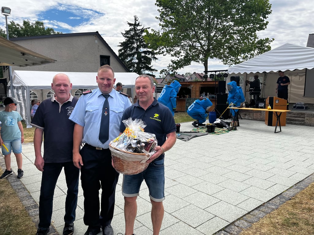 Mike Reinhold (l.) und Frank Deutschmann (r.) von der Feuerwehr Vitzenburg überreichten dem Weißenschirmbacher Wehrleiter David Seidenfaden einen Präsentkorb zum 90-jährigen Wehr-Jubiläum, während die Gefahrguteinheit des Saalekreises den Besuchern des Feuerwehrfestes eine Übung zeigte.