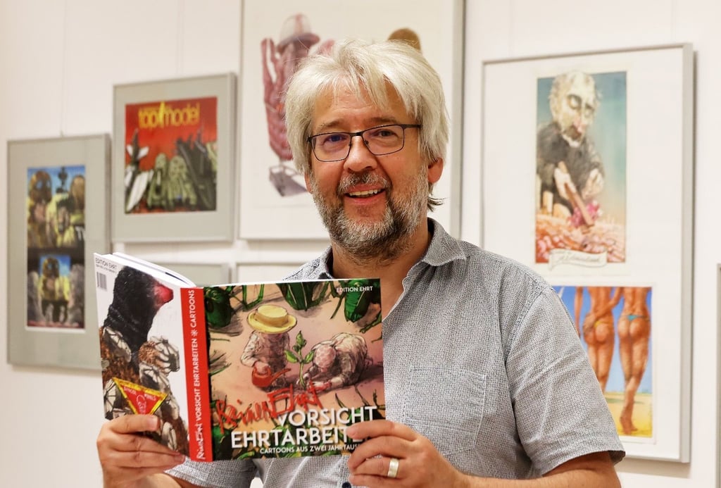 Die Ausstellung im Stadtarchiv zeigt Cartoons des Karikaturisten Rainer Erth. Stadtarchivar Ralf Jacob präsentiert den Katalog zur Schau.