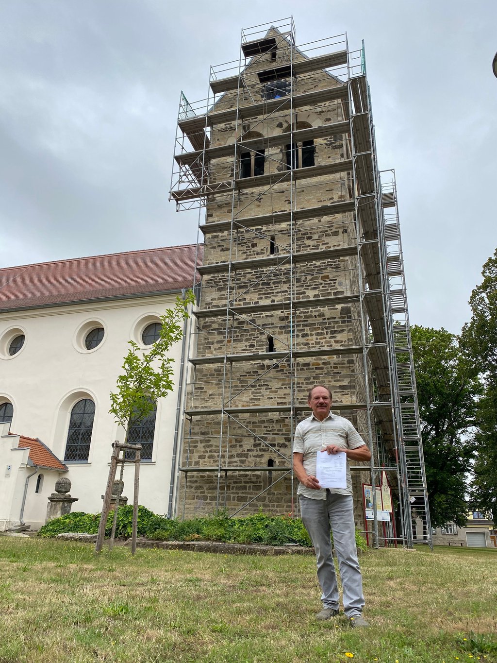 Ortsbürgermeister Hans-Jürgen Korn steht vor dem eingerüsteten Kirchturm und präsentiert stolz die Fördermittelbewilligung.
