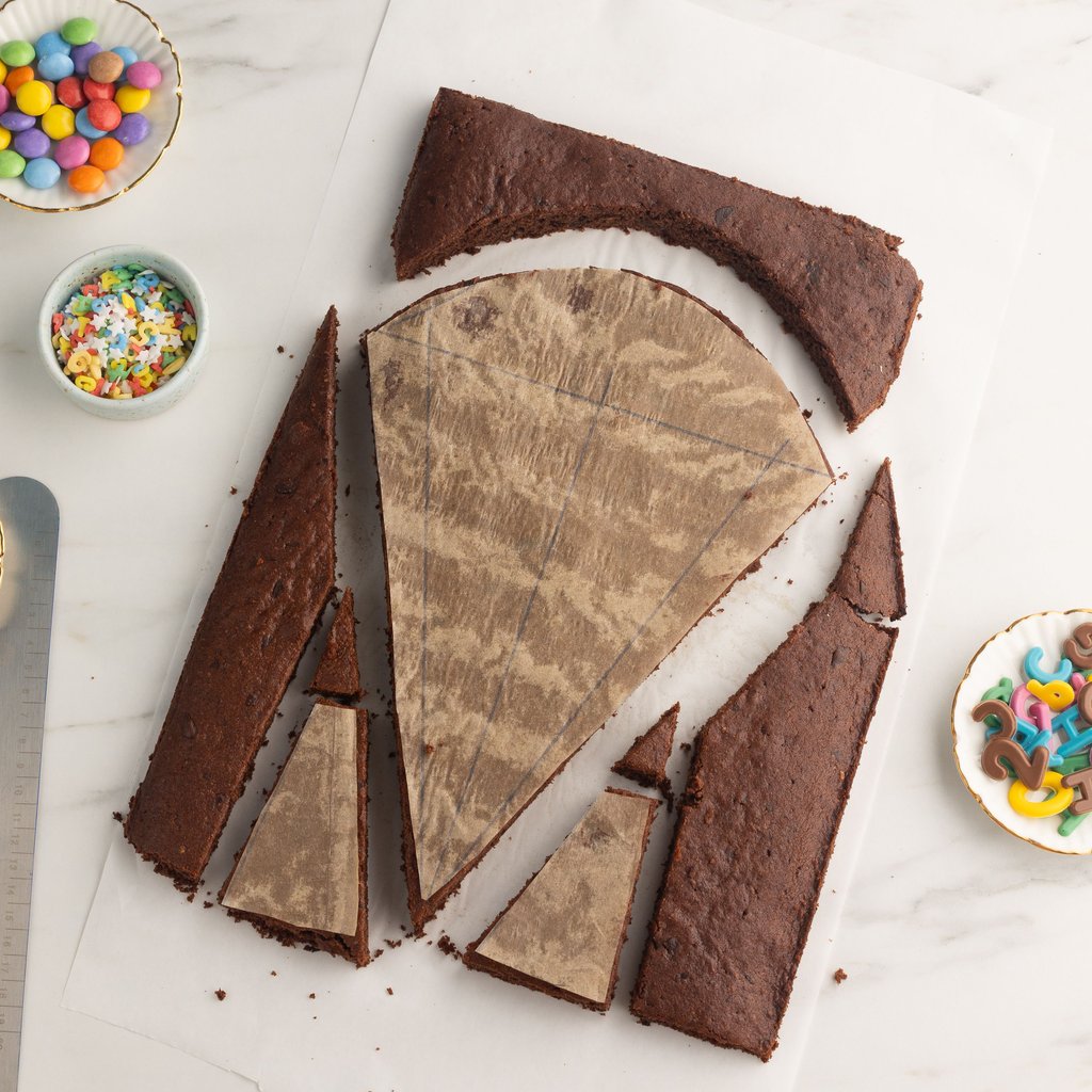 Ein klassischer Schokokuchen auf einem Blech mit Backrahmen ist die Basis für den Schultütenkuchen. Die zuvor gezeichneten Vorlagen auf Backpapier werden dann darauf gelegt und an den Rändern entlang zugeschnitten. Mutige können natürlich auch ohne Vorlage frei Hand zuschneiden.