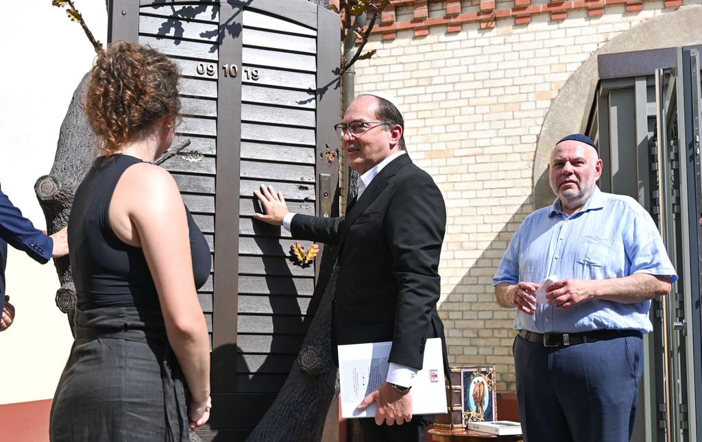 „Die Tür weist ja noch Einschusslöcher auf“: Bundesinnenminister Alexander Dobrindt (Mitte) im Gespräch mit der Künstlerin Lidia Edel. Der Gemeinde-Vorsitzende Max Privorozki (r.) hört zu.