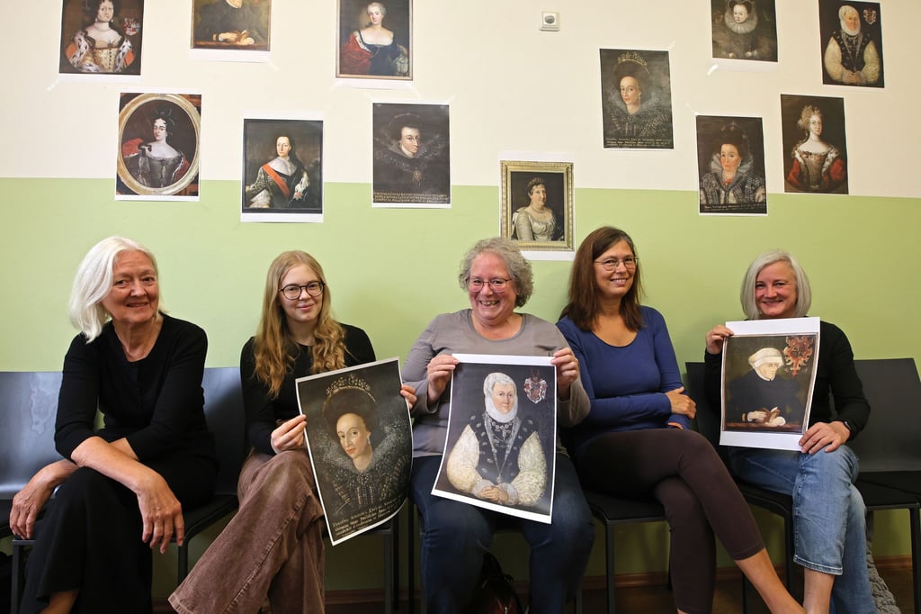 Teilnehmerinnen sitzen vor einem Fototermin mit historischen Porträts von ehemaligen Äbtissinnen im Stadtarchiv der Stadt Quedlinburg.