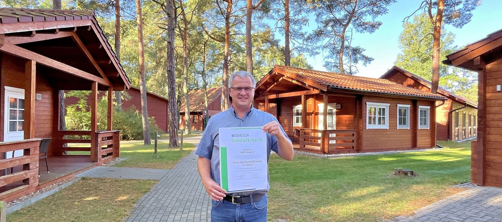 Feriendorf-Geschäftsführer Hendrik Steek posiert vor den Hotelzimmern der Anlage mit der Dehoga-Urkunde, die seinem Unternehmen Gold im Umweltcheck bescheinigt.