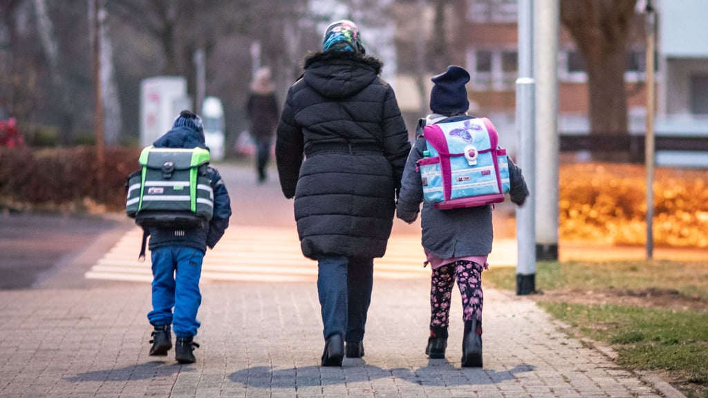 Zwei Schulkinder auf dem morgendlichen Schulweg – begleitet von einer erwachsenen Person. Es ist Herbst, die Straßen liegen noch im Dunkeln. Sicherheit auf dem Schulweg bleibt ein wichtiges Thema.