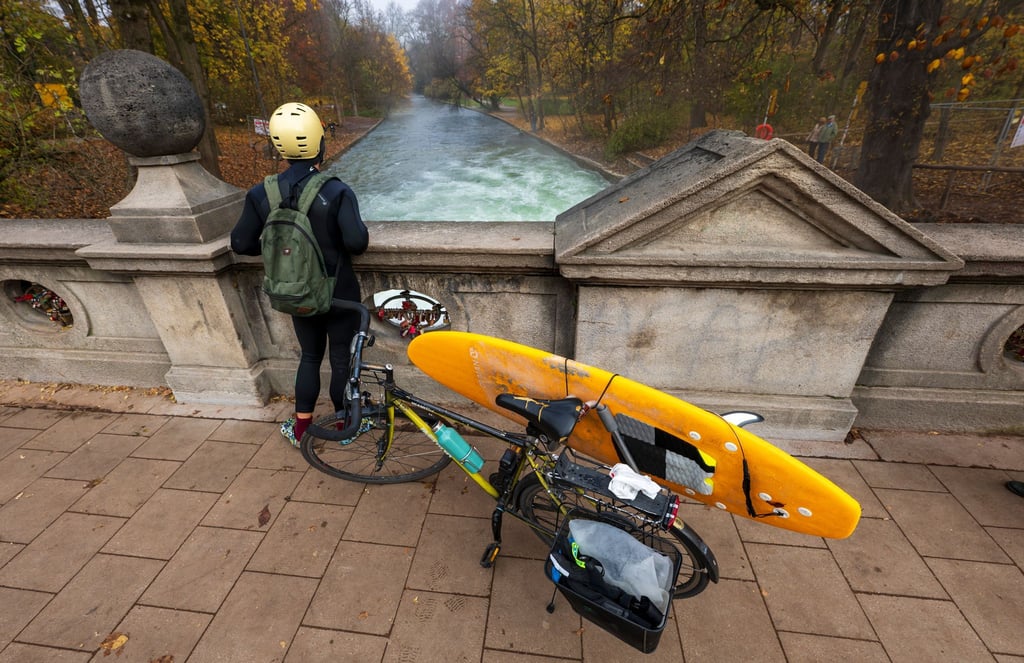 Ein Mann mit einem Surfboard an seinem Fahrrad schaut von einer Brücke auf die - nicht mehr vorhandene - Eisbachwelle im Englischen Garten.
