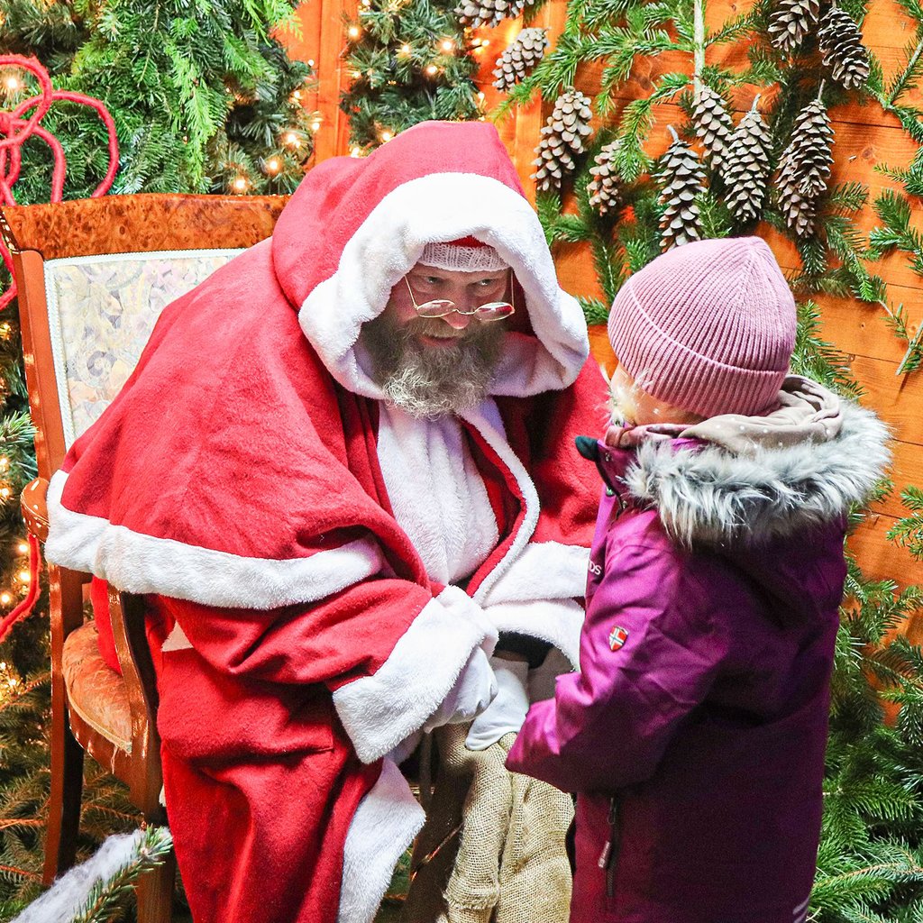 Ein als Weihnachtsmann verkleideter Mann sitzt mit einem Kind auf dem Schoß in einem festlich dekorierten Bereich des Weihnachtsmarktes Leuna 2024, umgeben von Tannengrün, Zapfen und weihnachtlicher Beleuchtung.  Weihnachtsmarkt, Leuna, Weihnachtsmann, Kind, Weihnachten, Nikolaus, Tannendekoration, Zapfen, 2024