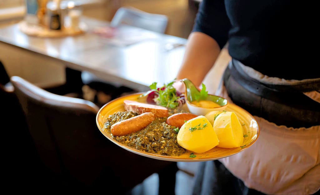 Eine Kellnerin serviert in einem Restaurant einen Teller mit Fleisch, Kartoffeln und Grünkohl. 