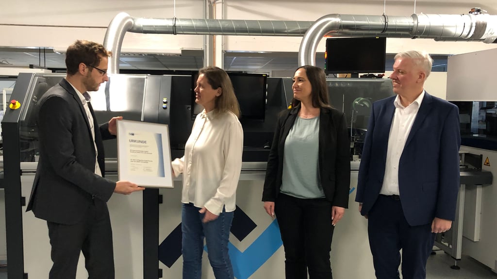 Vor der Selektivlötanlage übergibt IHK-Referent Lukas Thormann die Ehrenurkunde für 25 Jahre EMAS-Zertifizierung an Janet Haller, Umweltmanagementbeauftragte bei Rundfunk Gernrode, Sina Dreyer, Assistentin der Geschäftsführung,  und Geschäftsführer Marko Haller (v. l.)